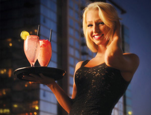 Smiling woman in a black dress holds a tray with two pink cocktails garnished with lemon, standing outdoors in front of a tall building at dusk.
