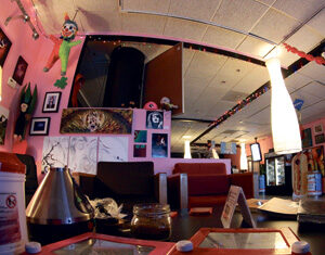 A cozy, colorful lounge with art-covered pink walls, Etch A Sketch toys on a table, comfy chairs, and soft lighting from tall lamps, creating a playful, relaxed atmosphere.