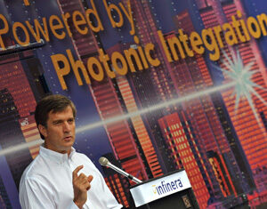 A man in a white shirt speaks at a podium with a microphone. Behind him is a colorful backdrop with cityscape graphics and the words Powered by Photonic Integration. The podium displays the Infinera logo.