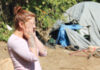 A woman with a bun and tattoos on her arms stands outdoors with her hands on her face. Behind her is a gray tent, tarps, and a bicycle in a wooded area, suggesting a campsite or encampment.