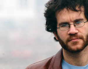 A man with curly dark hair, a beard, and glasses wears a brown leather jacket over a blue shirt with a graphic print. The background is blurred and outdoor.