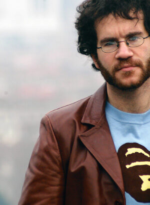 A man with curly dark hair, a beard, and glasses wears a brown leather jacket over a blue shirt with a graphic print. The background is blurred and outdoor.