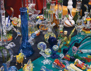 A variety of colorful glass pipes and bongs are displayed on a table, showcasing intricate designs and shapes in blues, reds, yellows, and greens. The pieces include animal figures and abstract forms.
