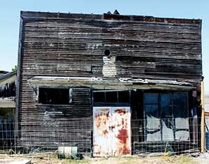 An old, weathered wooden building with peeling paint, rusted metal doors, broken windows, and a wire fence in front. The structure appears abandoned and neglected, with a clear blue sky overhead.