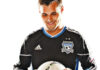 A soccer player wearing a black San Jose Earthquakes jersey looks down and smiles while holding a white soccer ball in both hands.