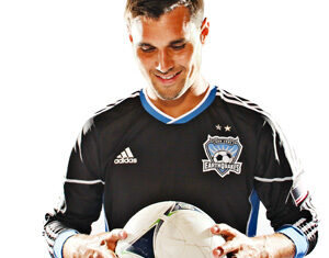 A soccer player wearing a black San Jose Earthquakes jersey looks down and smiles while holding a white soccer ball in both hands.