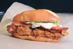 A crispy fried chicken sandwich with lettuce, tomato, and bacon on a toasted bun, placed on brown parchment paper.