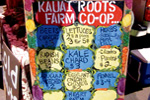 A colorful chalkboard sign at a farmers market reads Kauai Roots Farm Co-op and lists produce such as beets, lettuce, carrots, kale, chard, organic sweet corn, pineapple, and more with prices.