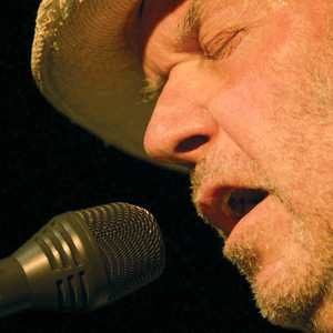Close-up of an older man with a beard and hat singing passionately into a microphone, eyes closed, against a dark background.