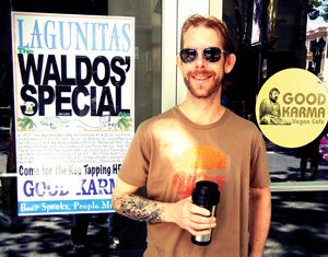 A man wearing sunglasses and holding a travel mug stands smiling outside Good Karma Vegan Cafe, next to a Lagunitas Waldo’s Special sign. Sunlight creates a reflection on his shirt and the glass door behind him.