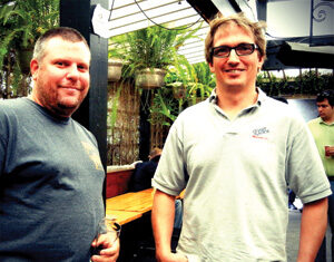 Two men are standing and smiling at the camera in an outdoor setting with plants and a wooden bench in the background. One man holds a drink while others are seen in the background.