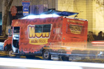 A red food truck with Wow and Club Truck written on the side is parked on a city street at night, with lights and blurred motion from passing cars in the foreground.