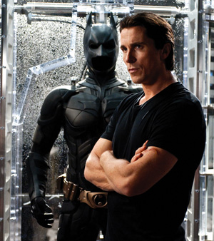 A man in a black t-shirt stands with arms crossed in front of a display case containing a black armored Batman suit with a utility belt.