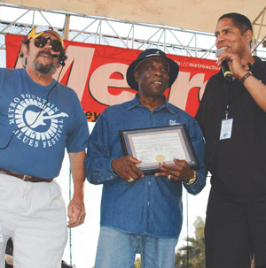 Three men stand on stage under a tent. The man in the middle holds a framed certificate and smiles, while the other two speak into microphones. A red Metro banner hangs in the background.