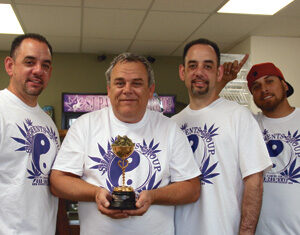 Four men wearing matching white T-shirts with a purple yin-yang logo stand together indoors. The man in the center is holding a trophy and smiling, while the others stand close, smiling at the camera.