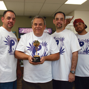 Four men wearing matching white T-shirts with a purple yin-yang logo stand together indoors. The man in the center is holding a trophy and smiling, while the others stand close, smiling at the camera.