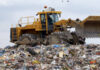 A yellow bulldozer sits on top of a large pile of mixed garbage and waste at a landfill site under a cloudy sky.