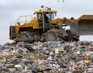 A yellow bulldozer sits on top of a large pile of mixed garbage and waste at a landfill site under a cloudy sky.