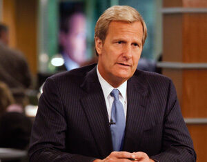 A man in a suit and tie sits at a news desk with papers and a notepad in front of him, appearing serious and focused in a newsroom setting.