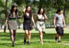 Four young adults walk together in a sunny park. Three women in dresses laugh and walk in front, while one person in a t-shirt and shorts walks a little behind them. Trees and green grass fill the background.
