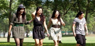 Four young adults walk together in a sunny park. Three women in dresses laugh and walk in front, while one person in a t-shirt and shorts walks a little behind them. Trees and green grass fill the background.