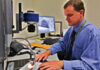 A man in a blue shirt sits at a desk using a computer and specialized equipment, with monitors displaying detailed images and documents around him.