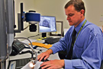A man in a blue shirt sits at a desk using a computer and specialized equipment, with monitors displaying detailed images and documents around him.
