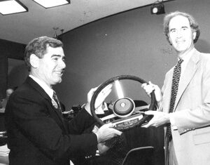 Two men in suits stand indoors, smiling as they hold a large car steering wheel together, possibly during a presentation or award ceremony. The setting appears to be a formal office or meeting room.