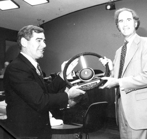 Two men in suits stand indoors, smiling as they hold a large car steering wheel together, possibly during a presentation or award ceremony. The setting appears to be a formal office or meeting room.