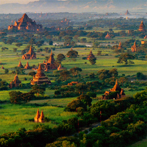 A lush green landscape dotted with ancient temples and pagodas, with distant mountains under a hazy sky, likely in Bagan, Myanmar.