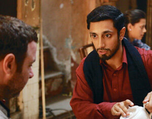 A man in a red shirt pours tea from a white teapot into a cup for another man at a patterned table in a rustic, warmly lit café. A woman sits in the background.
