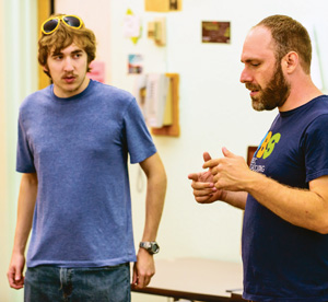 Two men stand indoors, facing each other. One wears a blue shirt and sunglasses on his head, looking at the other man, who is gesturing with his hands while speaking. A desk and wall items are in the background.