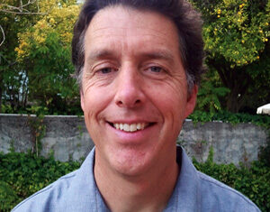 A man with short brown hair, wearing a light blue button-up shirt, smiles while standing outdoors in front of a stone wall and greenery.