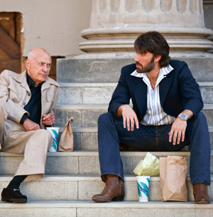 Two men sit on stone steps with drinks and paper bags beside them, talking. One man is older and dressed in a beige suit, while the other is younger, with a beard, wearing a dark jacket and jeans.