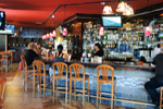 People sit on wooden chairs at a bar with colorful hanging lights, bottles displayed on shelves, and a bartender serving drinks. The bar has a relaxed atmosphere with a few patrons and TVs on the walls.