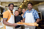 Five smiling men in a restaurant kitchen, two in front wearing aprons and holding a large dosa on a tray, with three others standing behind them, all appearing happy and friendly.