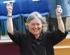 A smiling older woman with gray hair raises both hands in the air, standing outdoors in front of blue chairs and steps. She is wearing a black top and has glasses hanging from a cord around her neck.