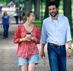 A woman in a red blouse and denim shorts holding a coffee cup walks and smiles at a man in a light blue shirt and jeans, who is holding a book, as they stroll along a tree-lined path.