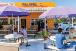 People sit at outdoor tables with purple umbrellas in front of a bright orange restaurant called Falafel, which offers shawarma, falafel, pizza, and burgers. Some patrons eat while others chat under a sunny sky.