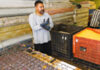 A man wearing gloves and a gray shirt stands beside large plastic crates in what appears to be an industrial or warehouse setting.