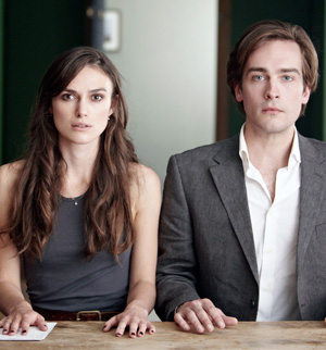 A woman with long brown hair and a sleeveless dark top sits next to a man in a gray blazer and white shirt at a wooden table, both looking directly at the camera with neutral expressions.