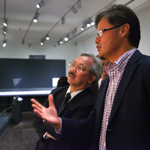 Two men in business attire stand in an art gallery or museum, engaged in conversation. One gestures with his hand while the other listens attentively, with exhibits visible in the background.