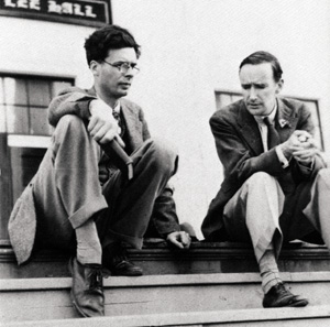 Two men in suits sit on outdoor steps, engaged in conversation. One man sits cross-legged holding an object, while the other leans forward with hands clasped. A building is visible in the background.