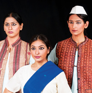 Three women stand together against a black background, wearing traditional Indian attire. Two wear embroidered vests, and one in front wears a white sari with a blue sash. The woman on the right wears a white cap.