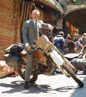 A man in a gray suit sits on a dusty motorcycle in a busy outdoor market with colorful carpets hanging behind him and people walking and sitting nearby.