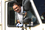 A smiling man wearing gloves leans out of the window of a white vehicle, looking at the camera.