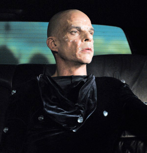 A bald person with pale skin, wearing a black outfit with shiny buttons, sits in the backseat of a car, looking thoughtfully to the side. The background is dimly lit.
