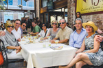 A group of eight adults sits together at an outdoor restaurant table, smiling at the camera. Plates, drinks, and sunglasses are on the table, and sunlight fills the lively, casual setting.