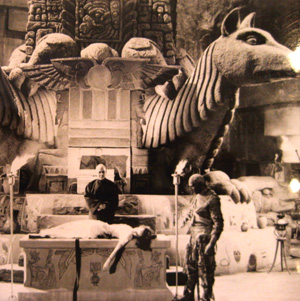 A black-and-white scene shows a woman lying on an altar, with two men near her—one appears to be a mummy. Behind them is a grand set featuring large winged statues and intricate carvings, evoking an ancient temple.