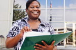 Smiling woman stands outside a building, holding a large green folder open in her hands. She wears a patterned blouse and looks confident and approachable.